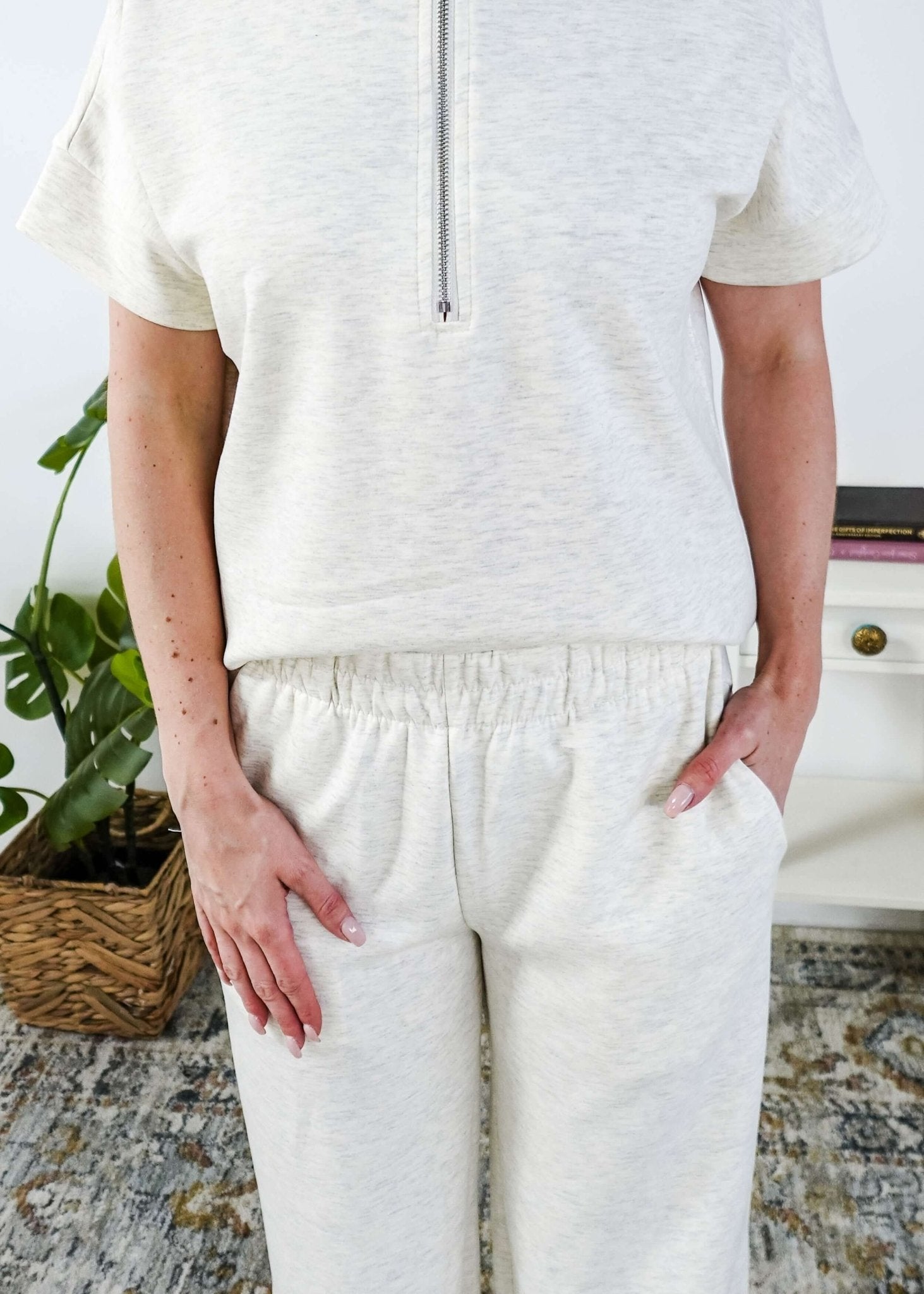Woman in a cream lounge outfit standing in a bedroom with a plant and bed in the background. Pants are an elastic pnt. 