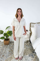 Woman in a cream lounge outfit standing in a bedroom with a plant and bed in the background