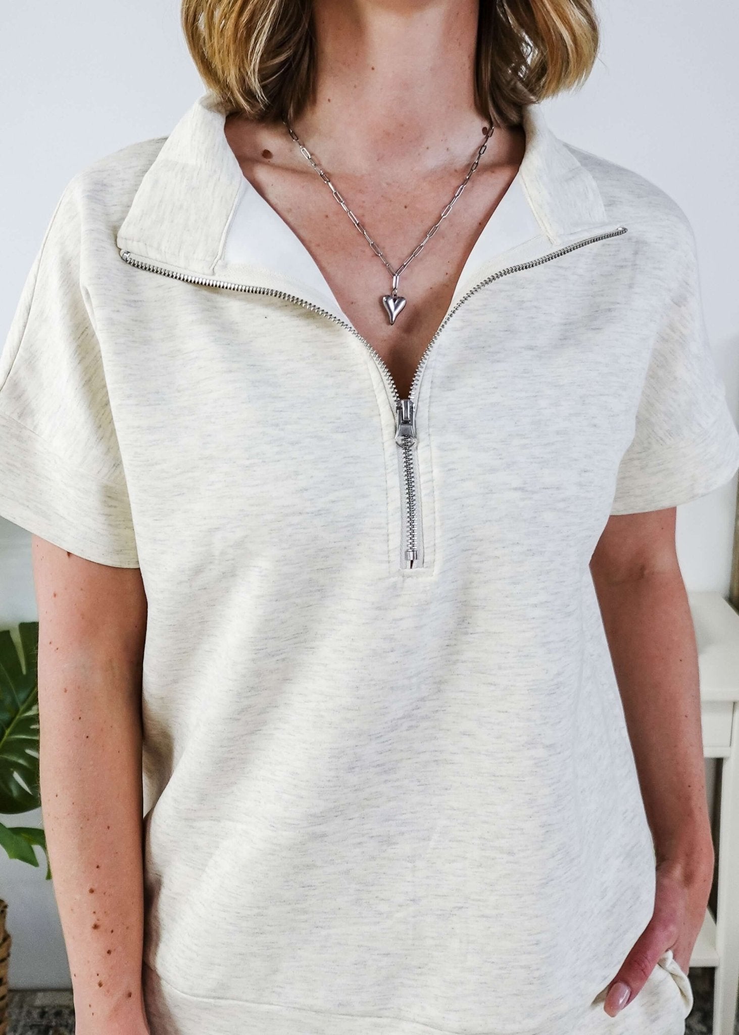 Woman in a cream lounge outfit standing in a bedroom with a plant and bed in the background. The sweater is a half zip top. 