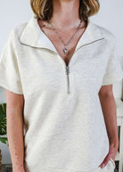 Woman in a cream lounge outfit standing in a bedroom with a plant and bed in the background. The sweater is a half zip top. 