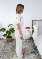 Woman in a cream lounge outfit standing in a bedroom with a plant and bed in the background