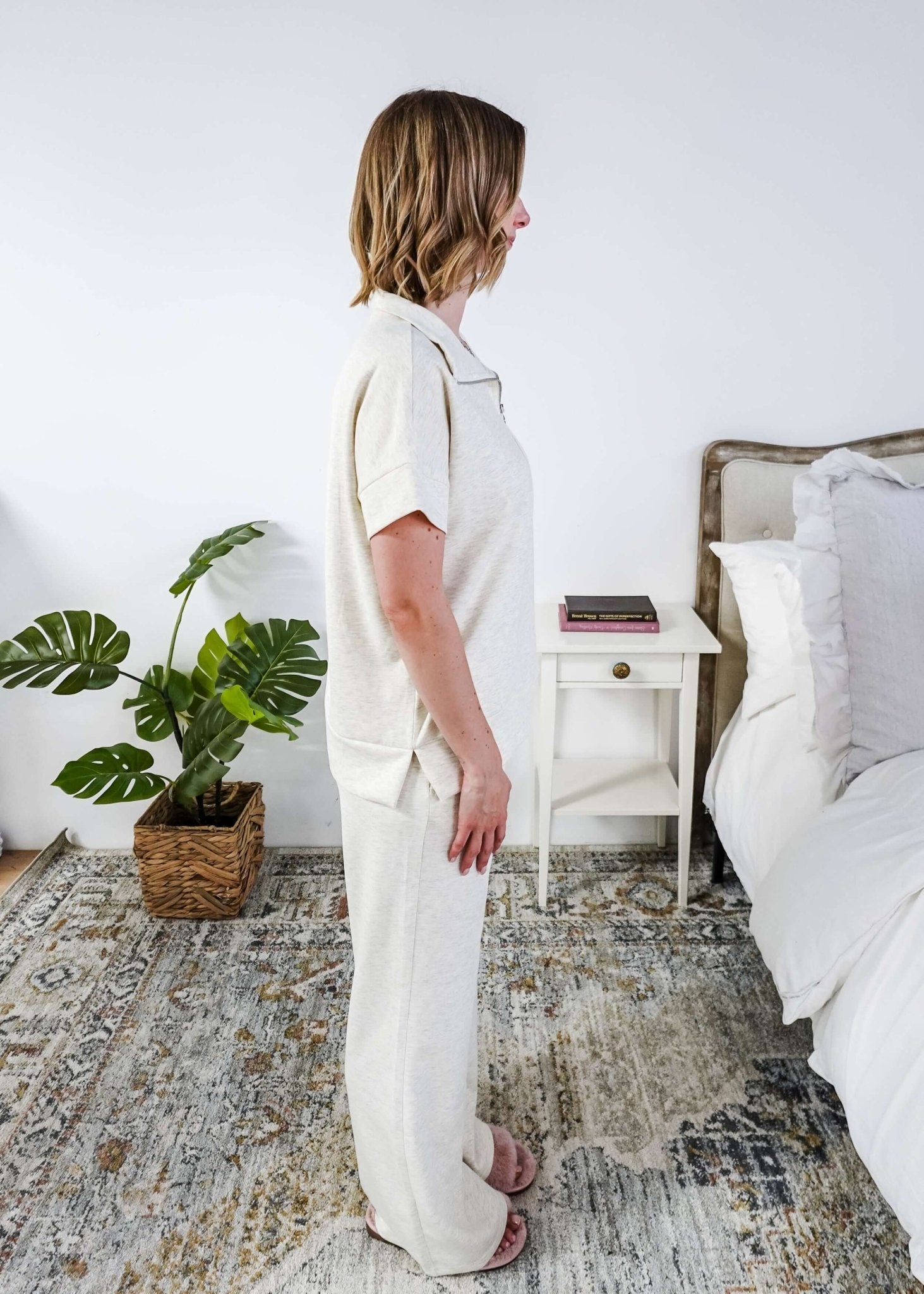 Woman in a cream lounge outfit standing in a bedroom with a plant and bed in the background