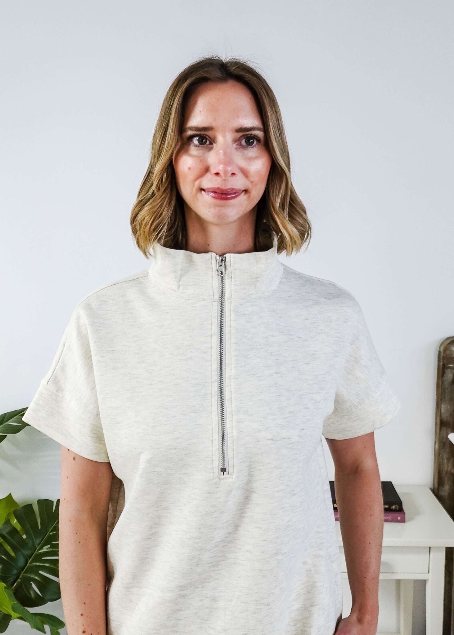 Woman in a cream lounge outfit standing in a bedroom with a plant and bed in the background. Sweater is zipped all the way up. 