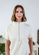 Woman in a cream lounge outfit standing in a bedroom with a plant and bed in the background. Sweater is zipped all the way up. 