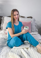 Woman in blue pajamas holding a mug on a bed with gray and pink pillows.