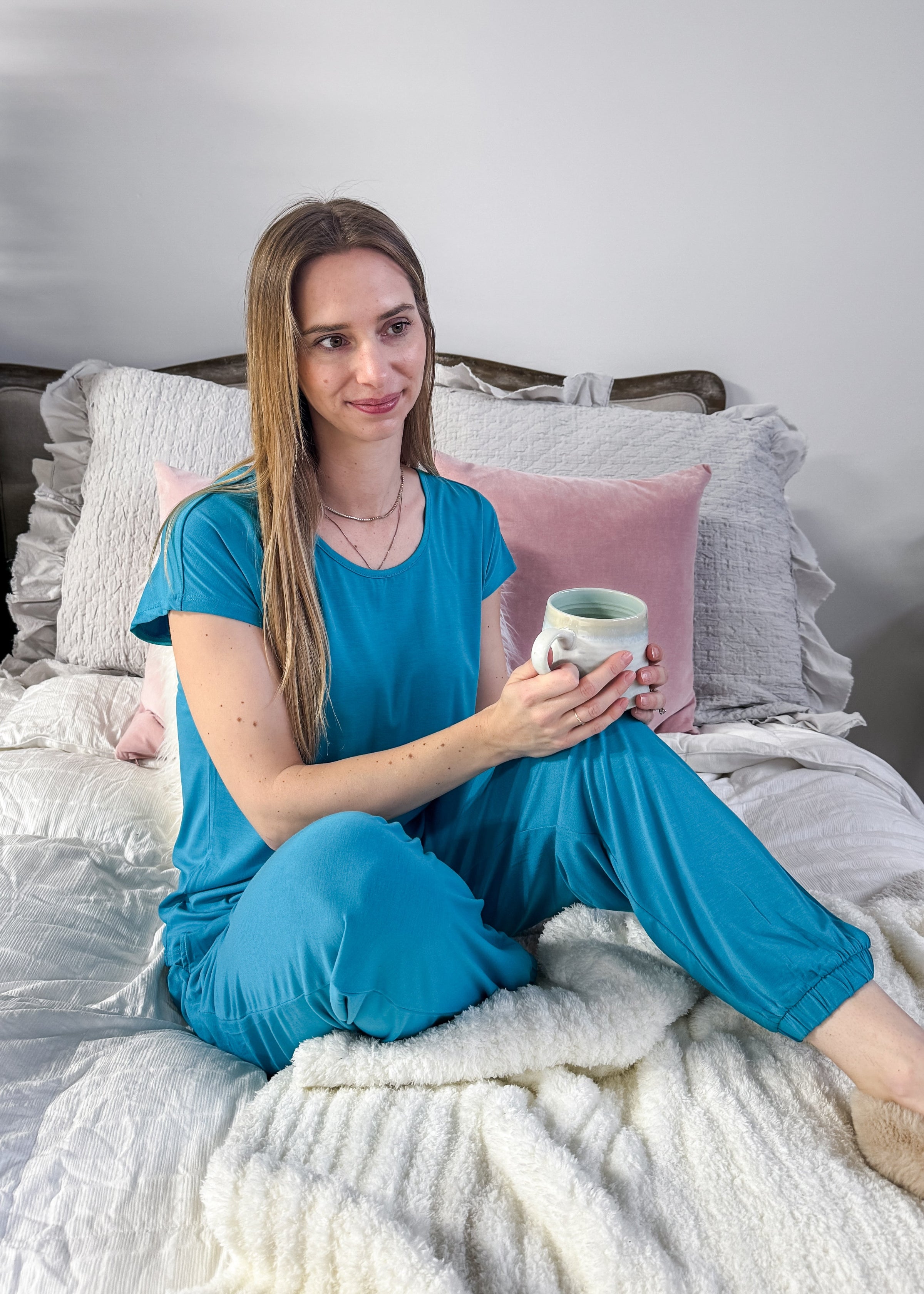 Woman in blue pajamas holding a mug on a bed with gray and pink pillows.