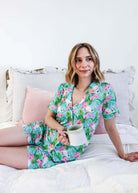 Woman enjoying a coffee in Tropic Like It's Hot Bamboo Short Sleeve Pajama Set by Georgia Rylan on a cozy bed.