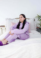 Woman in a purple outfit sitting on a bed with white bedding and gray pillows.