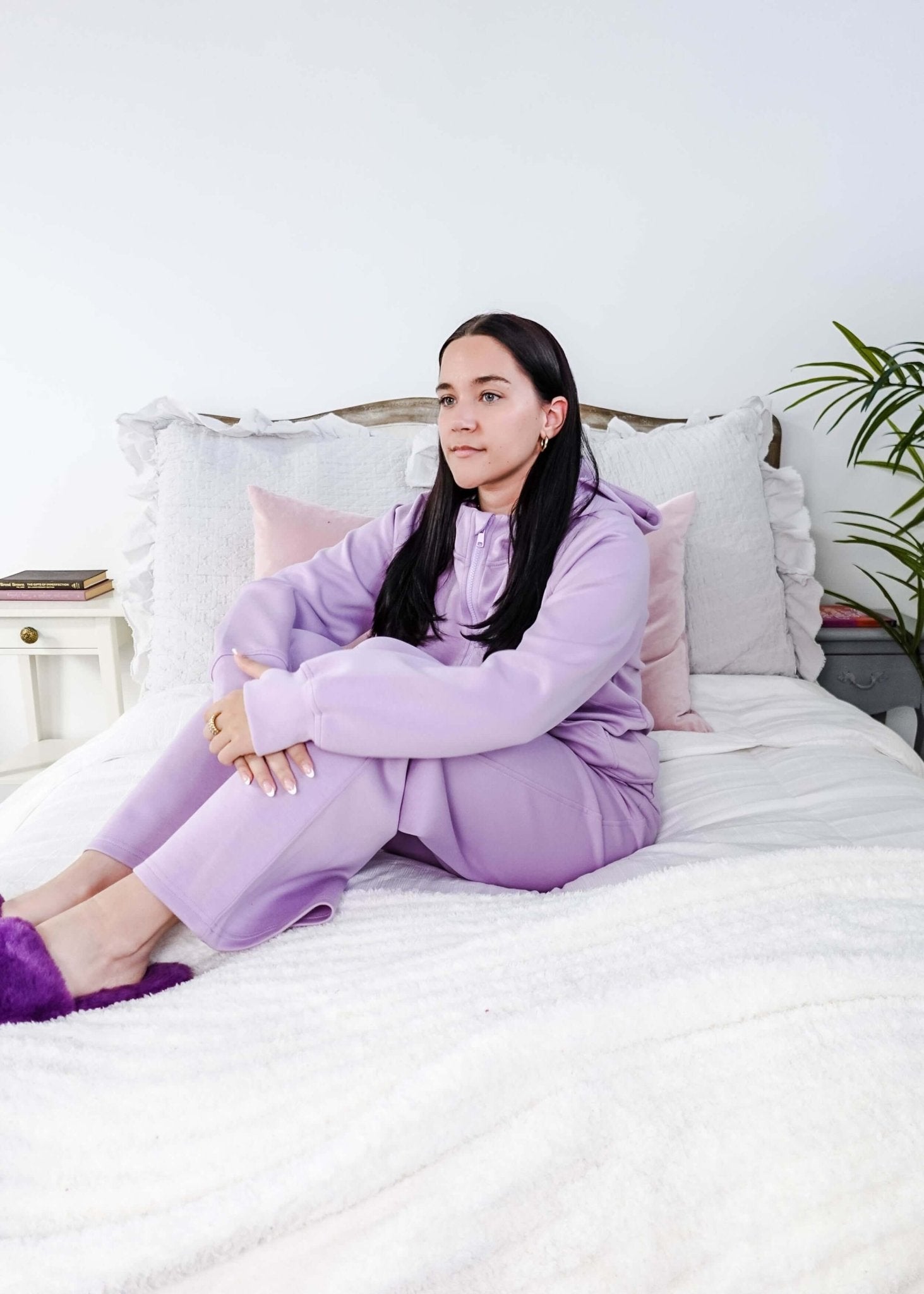 Woman in a purple outfit sitting on a bed with white bedding and gray pillows.